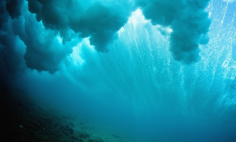 Deep blue ocean interior with light rays streaming down past the thick foam of a breaking wave overhead