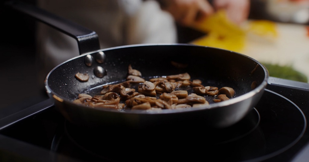 Close-up of sliced mushrooms cooking in a frying pan on an electric stovetop, with a person chopping vegetables in the background