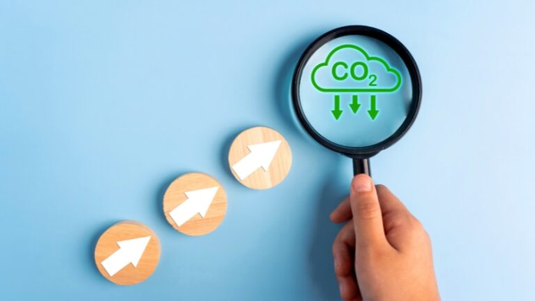 A hand holds a magnifying glass over a CO₂ cloud icon, symbolizing carbon flux tracking and monitoring