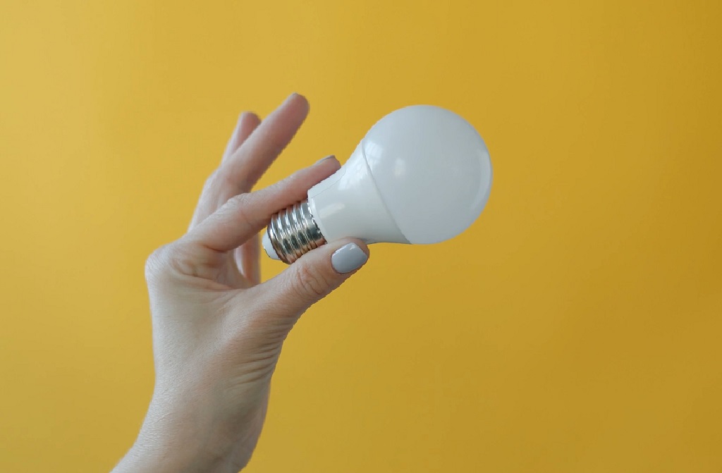 Hand holding an LED light bulb against a yellow background, symbolizing energy-saving choices