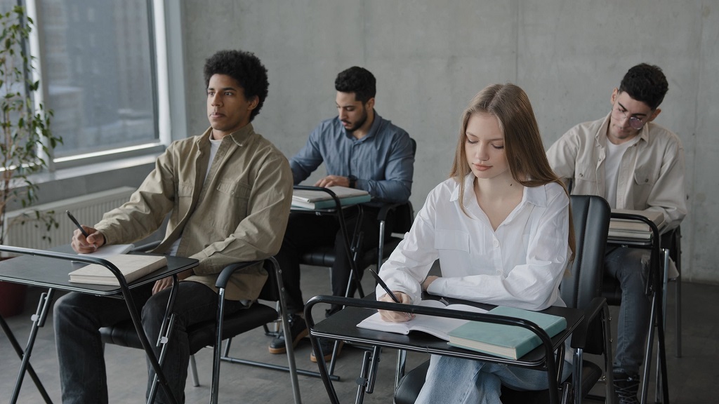 Group of students seated at desks in a classroom, focused on writing, with large windows allowing some natural light
