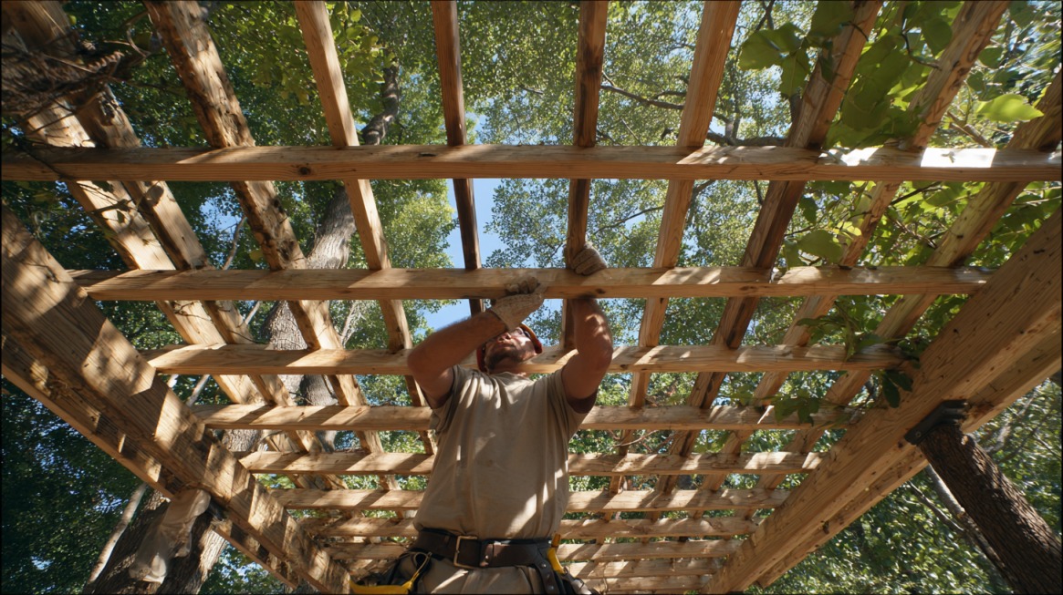 Man assembling a wooden pergola frame outdoors, viewed from below through wooden beams