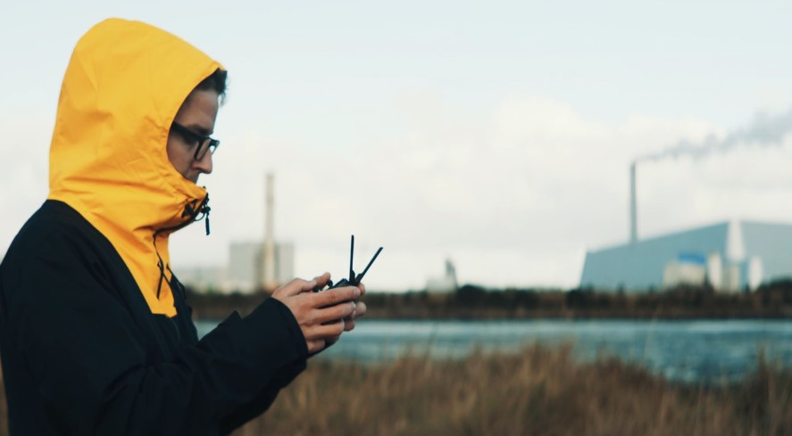 Drone pilot adjusts controls, a backdrop of industrial structures and a calm body of water