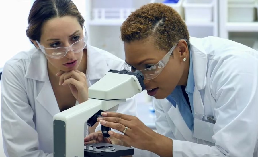 Two women in lab coats examining a microscope, engaged in scientific research and collaboration