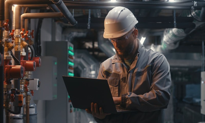 A man in a hard hat and work uniform examines a laptop in an industrial setting