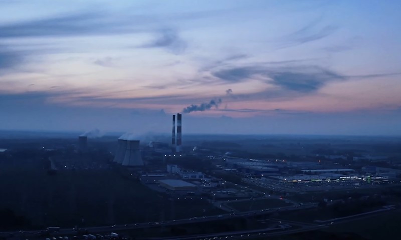A sprawling industrial plant with smokestacks emitting smoke is set against a dusk sky