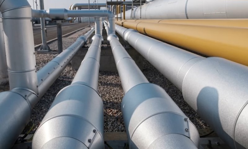 A series of large, silver and yellow industrial pipelines run parallel on a gravel path
