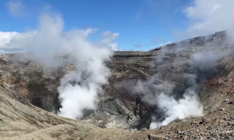 A volcanic crater emits white steam under a clear blue sky, surrounded by rough, rocky terrain
