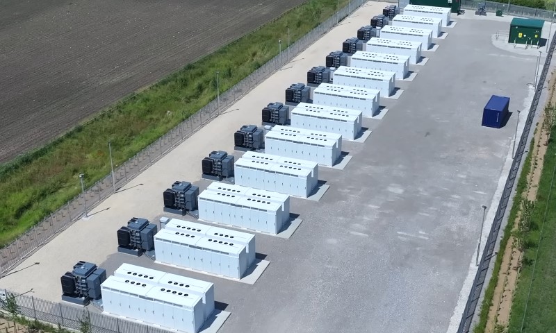 Aerial view of a grid-scale battery storage facility with rows of white containers and large cooling units on gravel