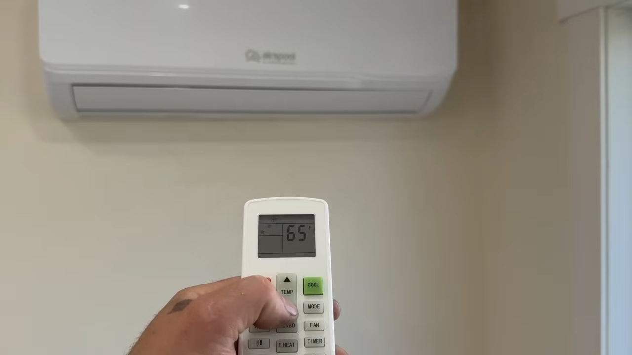 Hand holding a remote aimed at a wall mounted air conditioner showing 65°F.