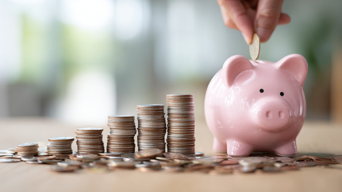 Hand placing a coin into a pink piggy bank beside stacks of coins arranged in increasing height