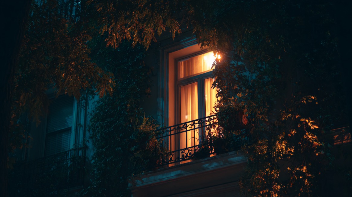 Warm light glowing from an open balcony window surrounded by leafy vines at night